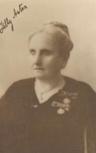 sepia photo of woman with her eyes closed. She has grey hair in a bun, black blouse, necklace and brooches. An ink autograph, "Tilly Aston", appears written diagonally across the upper left corner of the photograph